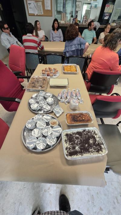 Una mesa cubierta con papel marrón muestra una gran variedad de platos caseros y postres, como bandejas de bizcochos, dulces envueltos y recipientes con tartas. Al fondo, varias personas están sentadas alrededor de otra mesa participando en la actividad desde una de nuestras agencias.