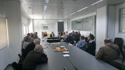 La imagen muestra a nuestros seniors reunidos, en la Sala Compromiso, en el CRE de Sevilla, alrededor de una gran mesa rectangular en un espacio moderno y bien iluminado. Sobre la mesa hay platos con aperitivos y bebidas, lo que indica un ambiente acogedor y participativo. Al fondo se aprecia un cartel con el logotipo de SUPO, reforzando el contexto del evento..