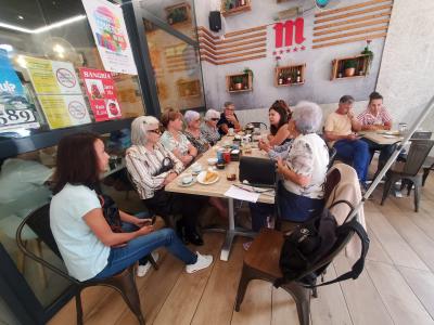 Grupo de socios compartiendo comida y conversación en un restaurante acogedor. Ambiente cálido, decoración con plantas y una gran letra “M” roja en la pared. Espíritu de comunidad y cercanía.
