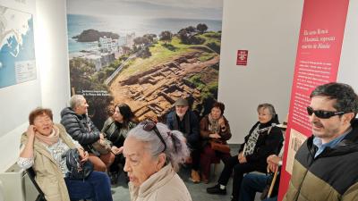 Fotografia que muestra distintos participantes durante la visita a Blandae. Se observa en el fondo una fotografía del antiguo asentamiento romano. 