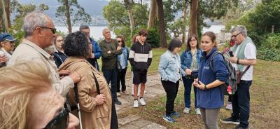 Personas participando en una visita guiada al aire libre, en un entorno natural junto al mar, con árboles, senderos de piedra y colinas al fondo.