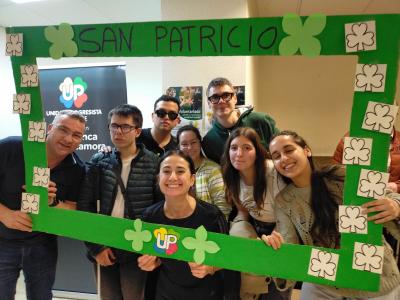 Un grupo de personas colocadas una al lado de otra, de pie, mirando hacia el frente. Entre todas sostienen un photocall, el cual está cubierto de tréboles haciendo referencia al Día de San Patricio. Entre los tréboles también aparece el logo de UP, integrado en el diseño del photocall.