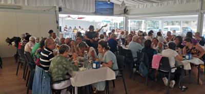 Personas reunidas en un comedor amplio y luminoso, sentadas alrededor de mesas compartiendo comida y conversación durante la sardiñada.