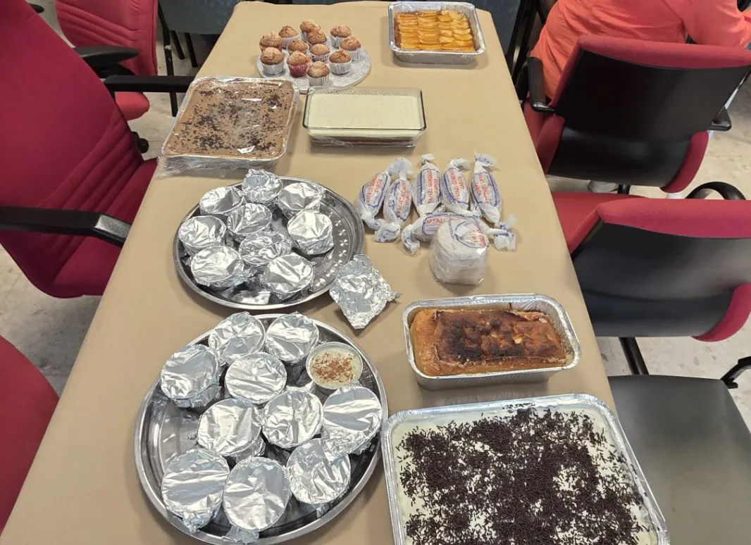 Una mesa cubierta con papel marrón muestra una gran variedad de platos caseros y postres, como bandejas de bizcochos, dulces envueltos y recipientes con tartas. Al fondo, varias personas están sentadas alrededor de otra mesa participando en la actividad desde una de nuestras agencias.