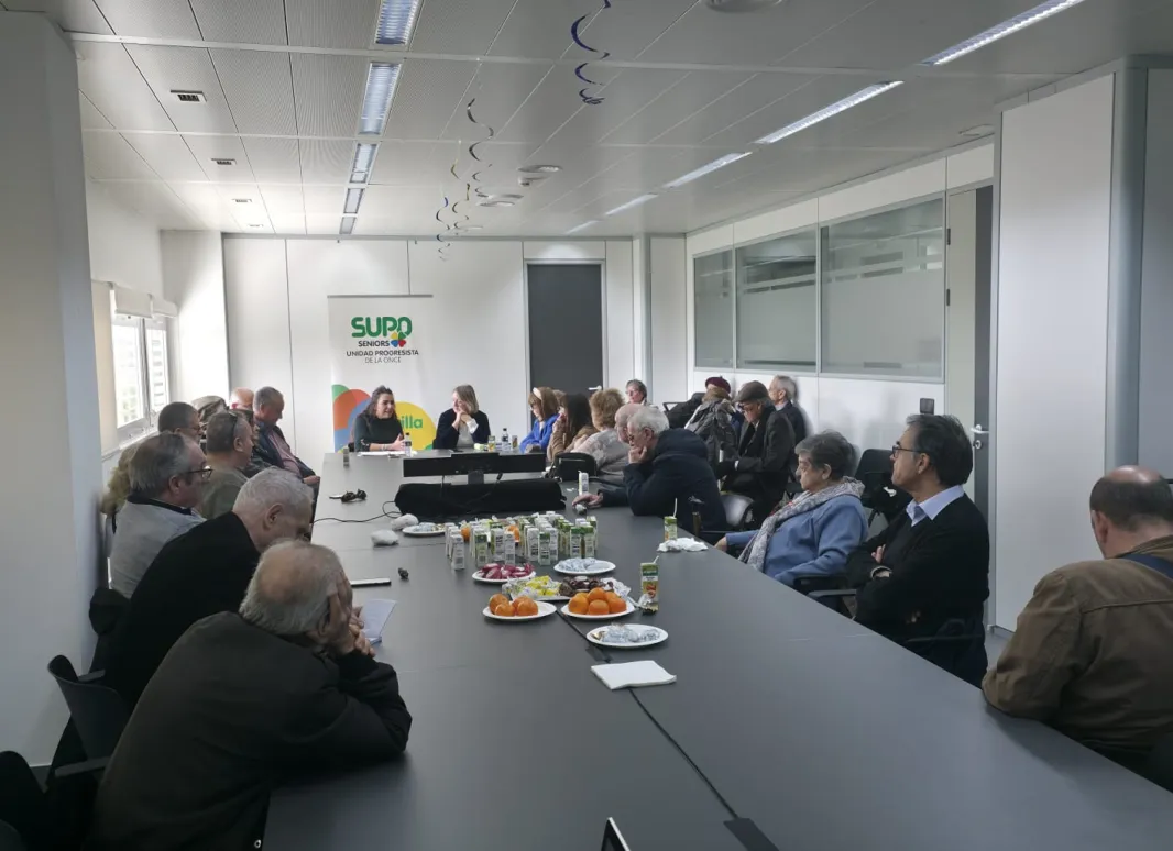 La imagen muestra a nuestros seniors reunidos, en la Sala Compromiso, en el CRE de Sevilla, alrededor de una gran mesa rectangular en un espacio moderno y bien iluminado. Sobre la mesa hay platos con aperitivos y bebidas, lo que indica un ambiente acogedor y participativo. Al fondo se aprecia un cartel con el logotipo de SUPO, reforzando el contexto del evento..