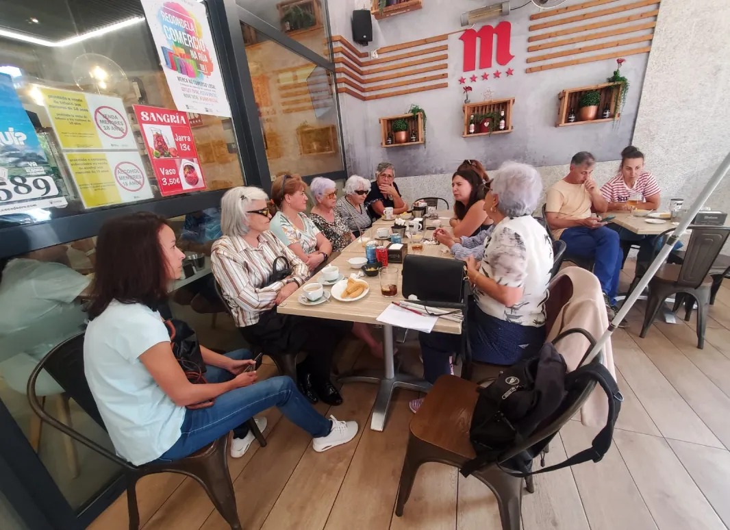 Grupo de socios compartiendo comida y conversación en un restaurante acogedor. Ambiente cálido, decoración con plantas y una gran letra “M” roja en la pared. Espíritu de comunidad y cercanía.