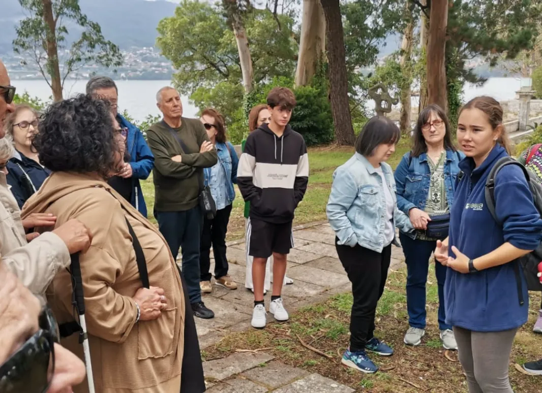 Personas participando en una visita guiada al aire libre, en un entorno natural junto al mar, con árboles, senderos de piedra y colinas al fondo.