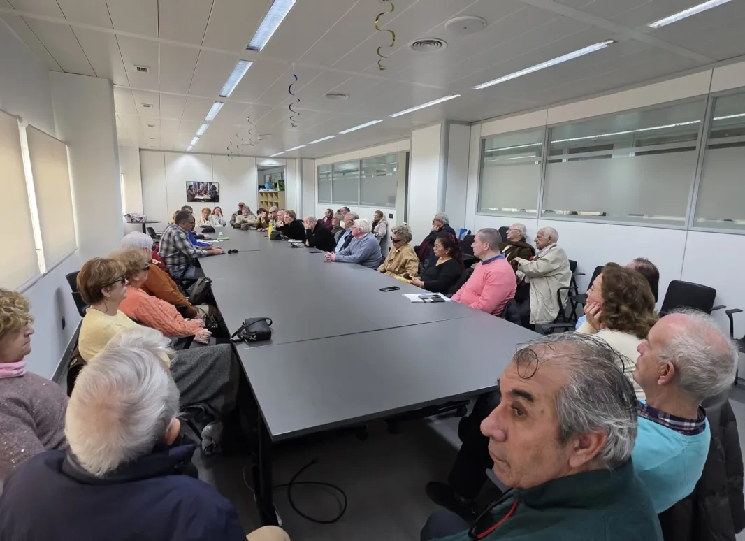 En la imagen se observa el desarrollo de la Asamblea de SUPO en Sevilla en una sala amplia y luminosa, dispuesta para reuniones de trabajo. Los asistentes ocupan los asientos alrededor de una gran mesa rectangular, lo que favorece un ambiente de diálogo y participación directa entre todas las personas presentes. El encuentro reúne a un grupo numeroso de socios y simpatizantes, sentados de forma ordenada, escuchando con atención las intervenciones. Sobre la mesa se aprecian documentos, libretas y algunos dis