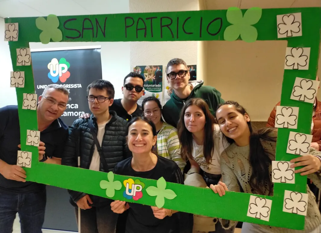 Un grupo de personas colocadas una al lado de otra, de pie, mirando hacia el frente. Entre todas sostienen un photocall, el cual está cubierto de tréboles haciendo referencia al Día de San Patricio. Entre los tréboles también aparece el logo de UP, integrado en el diseño del photocall.