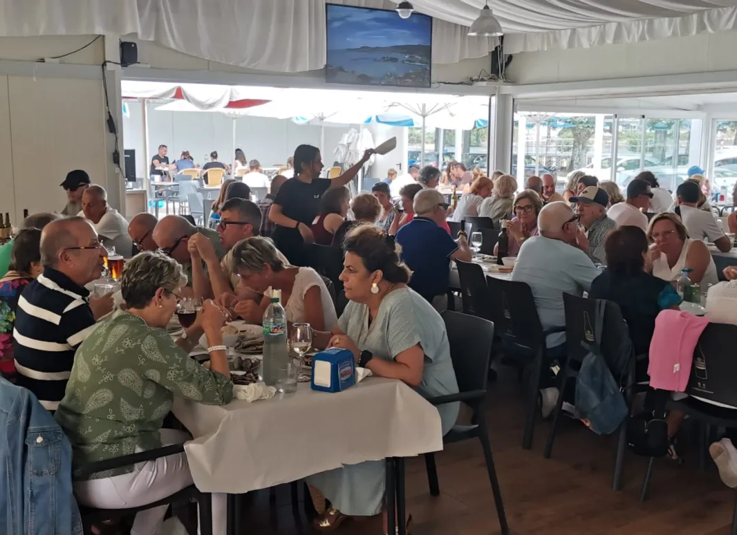 Personas reunidas en un comedor amplio y luminoso, sentadas alrededor de mesas compartiendo comida y conversación durante la sardiñada.