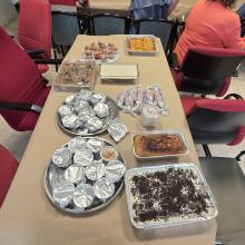 Una mesa cubierta con papel marrón muestra una gran variedad de platos caseros y postres, como bandejas de bizcochos, dulces envueltos y recipientes con tartas. Al fondo, varias personas están sentadas alrededor de otra mesa participando en la actividad desde una de nuestras agencias.