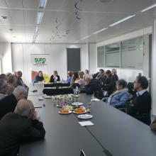 La imagen muestra a nuestros seniors reunidos, en la Sala Compromiso, en el CRE de Sevilla, alrededor de una gran mesa rectangular en un espacio moderno y bien iluminado. Sobre la mesa hay platos con aperitivos y bebidas, lo que indica un ambiente acogedor y participativo. Al fondo se aprecia un cartel con el logotipo de SUPO, reforzando el contexto del evento..