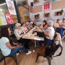 Grupo de socios compartiendo comida y conversación en un restaurante acogedor. Ambiente cálido, decoración con plantas y una gran letra “M” roja en la pared. Espíritu de comunidad y cercanía.