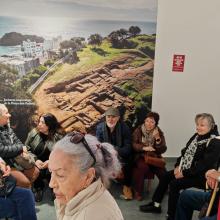Fotografia que muestra distintos participantes durante la visita a Blandae. Se observa en el fondo una fotografía del antiguo asentamiento romano. 