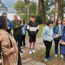 Personas participando en una visita guiada al aire libre, en un entorno natural junto al mar, con árboles, senderos de piedra y colinas al fondo.