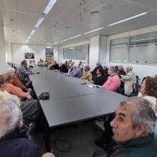 En la imagen se observa el desarrollo de la Asamblea de SUPO en Sevilla en una sala amplia y luminosa, dispuesta para reuniones de trabajo. Los asistentes ocupan los asientos alrededor de una gran mesa rectangular, lo que favorece un ambiente de diálogo y participación directa entre todas las personas presentes. El encuentro reúne a un grupo numeroso de socios y simpatizantes, sentados de forma ordenada, escuchando con atención las intervenciones. Sobre la mesa se aprecian documentos, libretas y algunos dis