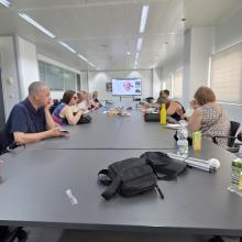 La imagen muestra a un grupo de personas reunidas en una sala del CRE de Sevilla, sentadas alrededor de una mesa y siguiendo en una pantalla una ponencia en formato videoconferencia, en el marco del webinar sobre la “Lucha contra la Soledad No Deseada”. La disposición híbrida, con participantes presenciales y conexión online, refleja un encuentro participativo en el que los asistentes comparten un espacio de atención y trabajo conjunto, acorde con la actividad de reflexión y sensibilización desarrollada por