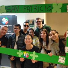 Un grupo de personas colocadas una al lado de otra, de pie, mirando hacia el frente. Entre todas sostienen un photocall, el cual está cubierto de tréboles haciendo referencia al Día de San Patricio. Entre los tréboles también aparece el logo de UP, integrado en el diseño del photocall.