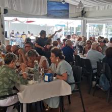 Personas reunidas en un comedor amplio y luminoso, sentadas alrededor de mesas compartiendo comida y conversación durante la sardiñada.